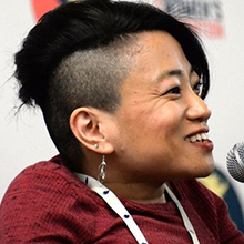 Mia Ives-Rublee sits in front of a microphone, smiling. She has short dark hair in a modern cut and is wearing a red shirt. 